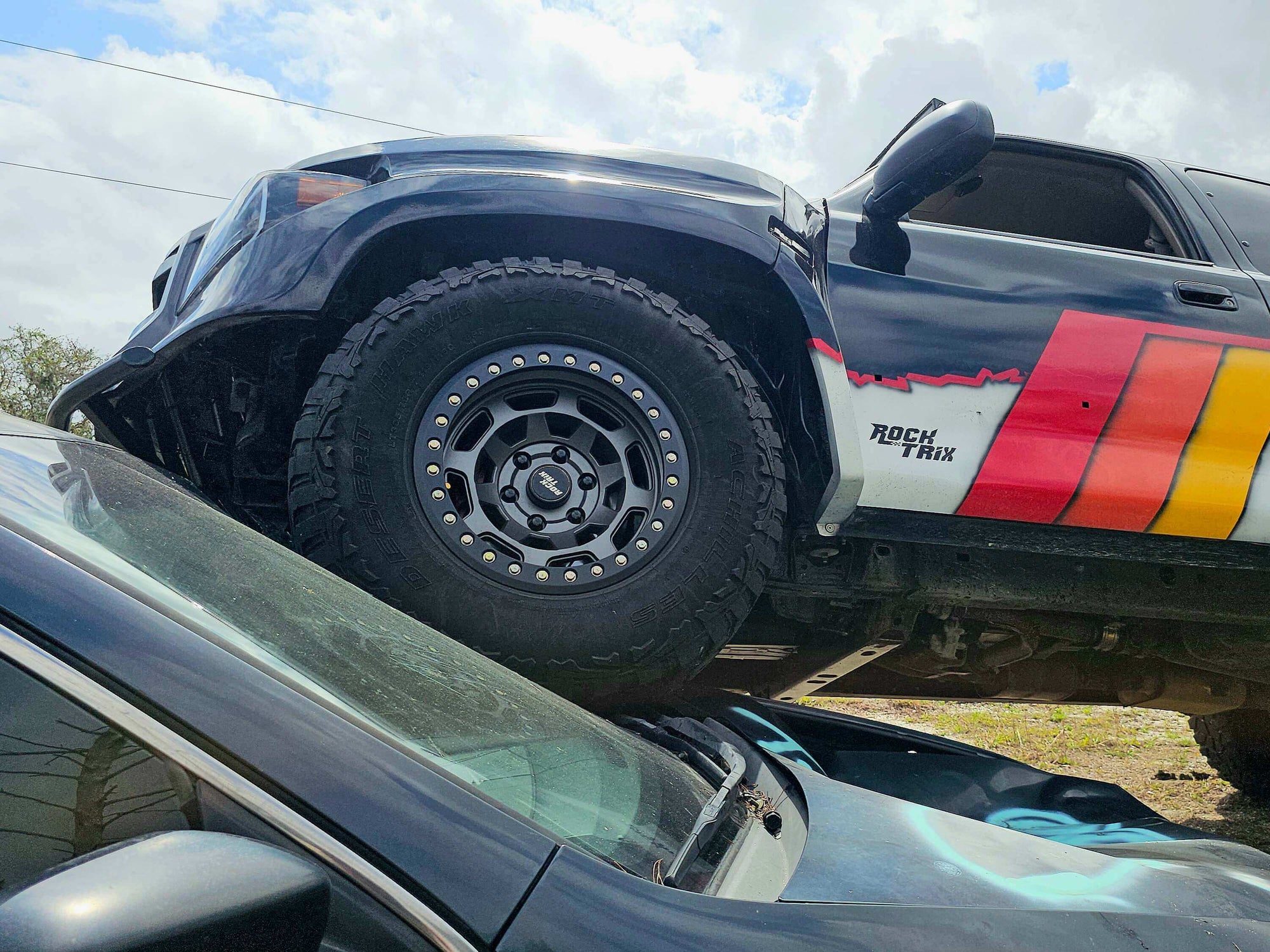 Toyota prerunner off-road vehicle with large tire and 'Rock Trix' branding on a cloudy day running over a junk car.